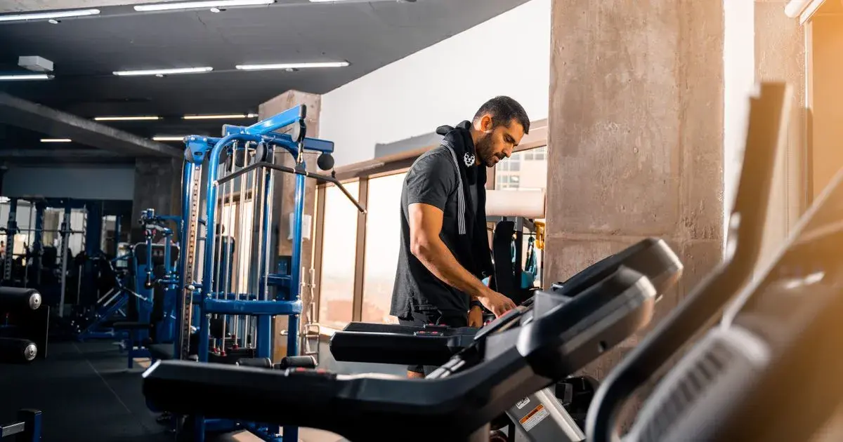 A man with a towel over his shoulders setting up a treadmill in a gym, part of a routine to stay consistent at the gym