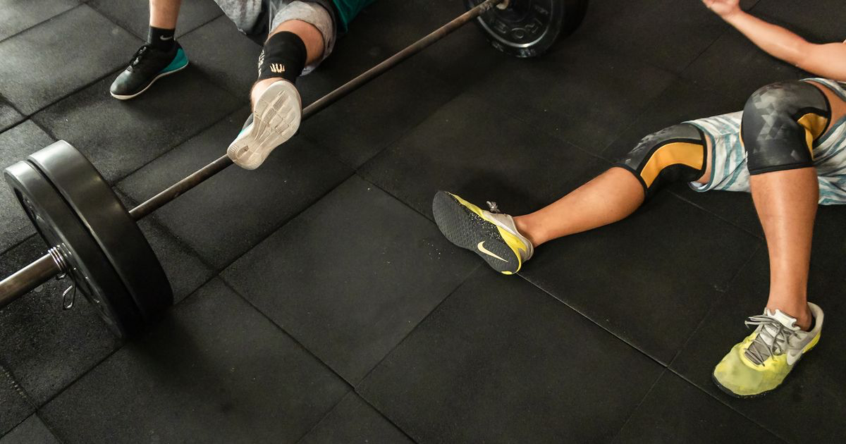 Two lifters resting on a gym floor beside a loaded barbell after a training session