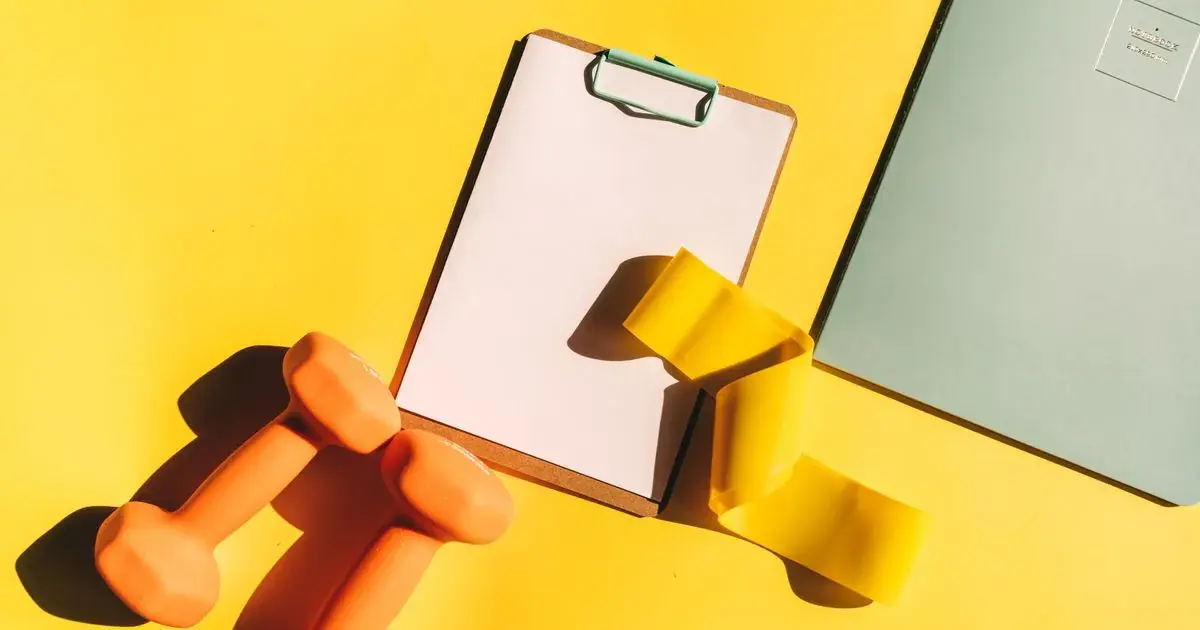 Workout log template on a clipboard beside dumbbells and a notebook
